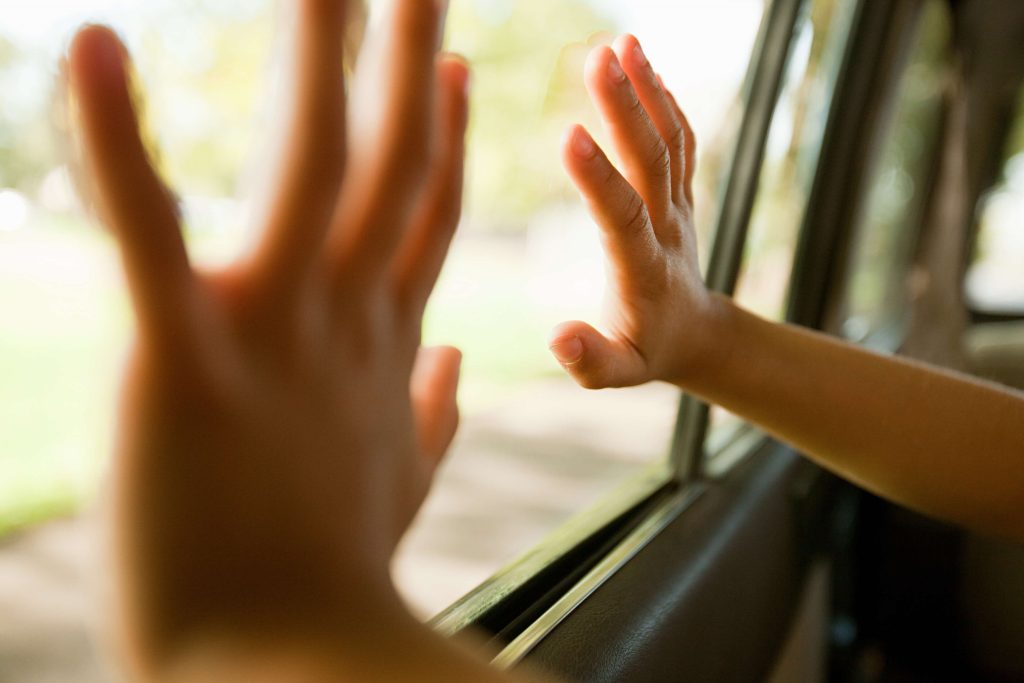 An infant is pictured here placing hands on a car set windown, the bright sun shining in the background.