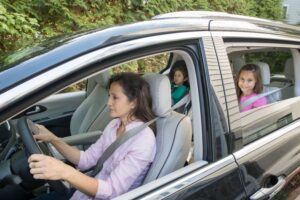 A mom is pictured here transporting two kids in a passenger vehicle. All three are buckled up.