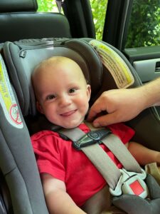 An infant smiles and sticks out his tongue while being secured in his car seat.