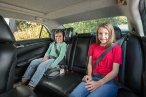 A little boys is pictured here sitting in a booster seat. And a little girl is fastened in with a seat belt.