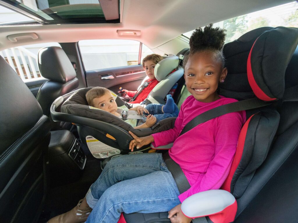 A school-aged child is pictured here smiling while riding in a booster seat. Two friends are pictured next to her in the back seat.