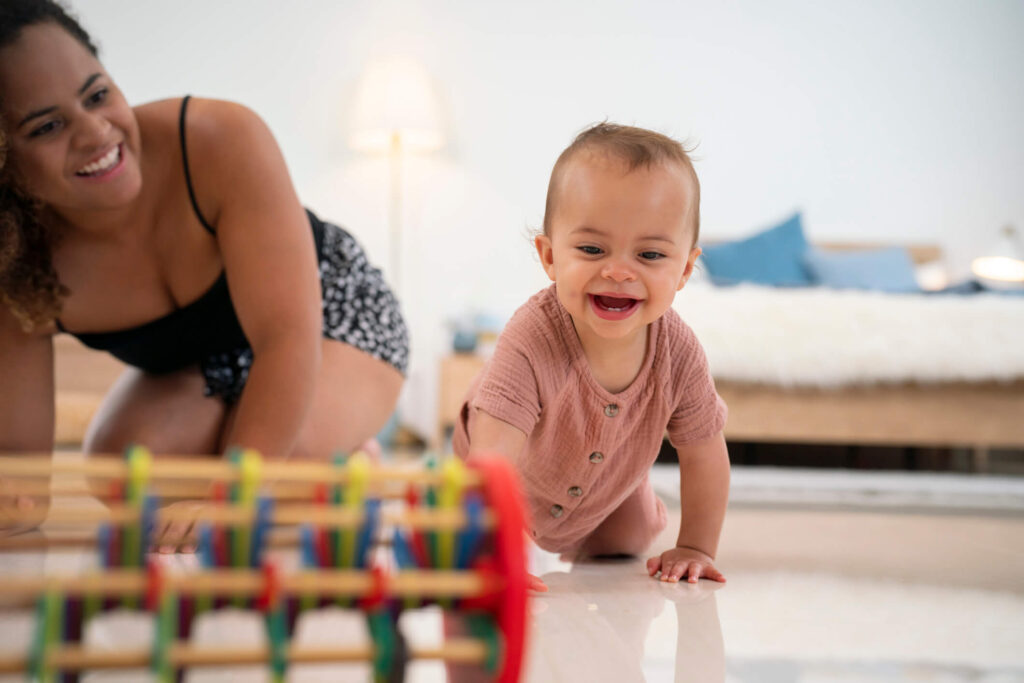 A mother kneels on the floor and smiles as she watches her happy infant crawl toward a toy.
